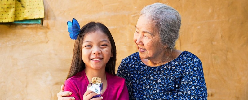 smiling girl eats a granola bar with a blue butterfly in her hair while an older woman smiles with her arm around the girl's shoulder
